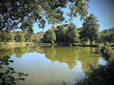 L’étang des Gentianes sous les arbres ; au-dessus de la rive, le toit de l’église Saint-Nicolas
